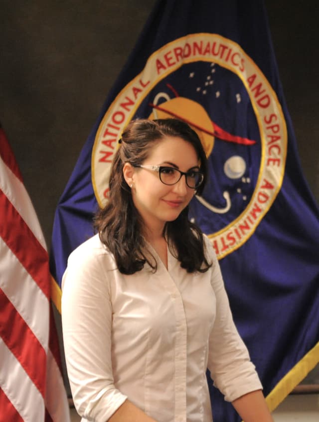 A professional in a white collared shirt with brown hair, sitting in front of the US NASA's flags.